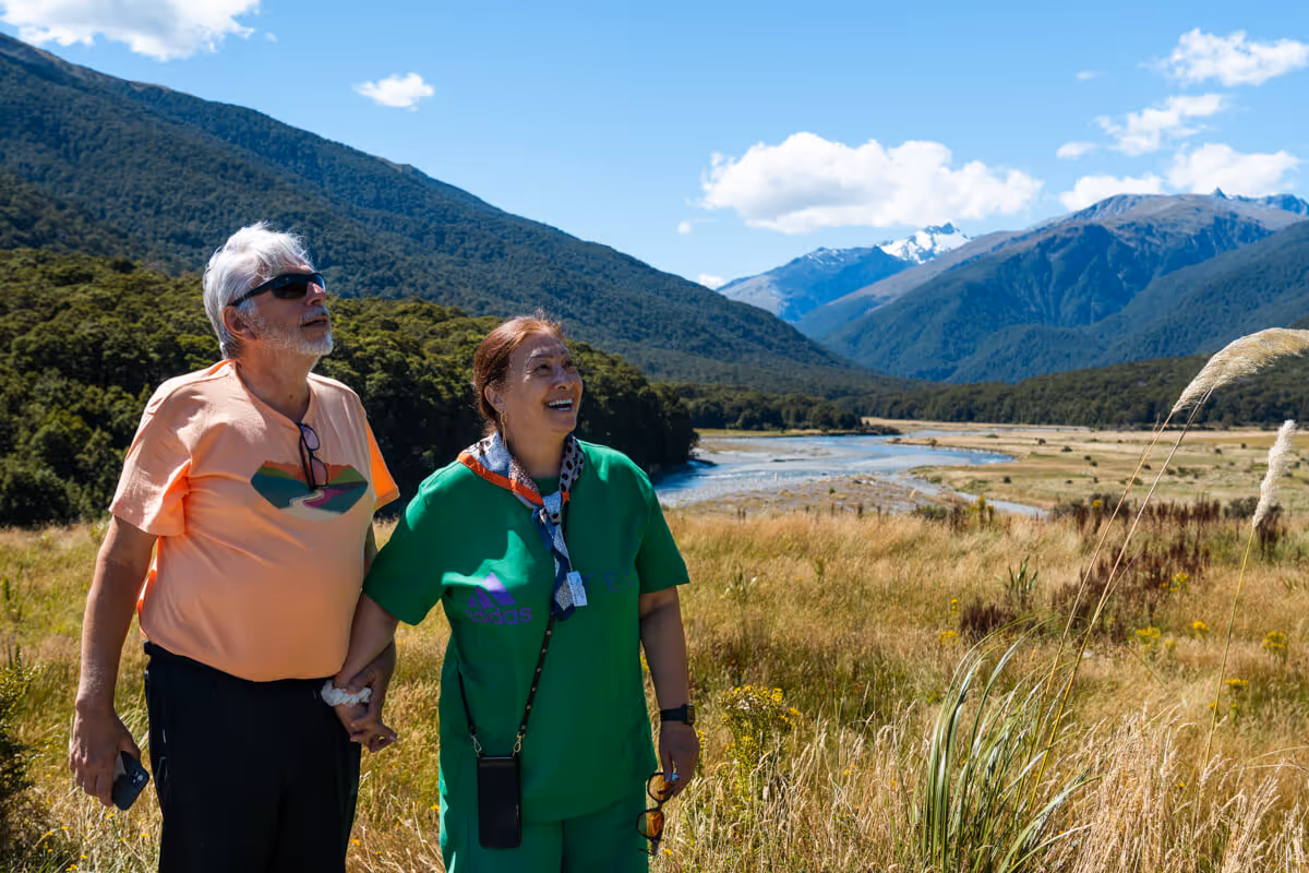 Older couple holding hands and smiling while standing in a grassy field with mountains and a river in the background under a blue sky.
