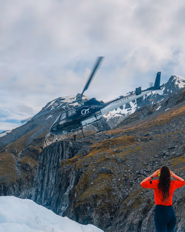 Blue helicopter flying near rocky snowy mountains with a person in a red jacket watching from below.