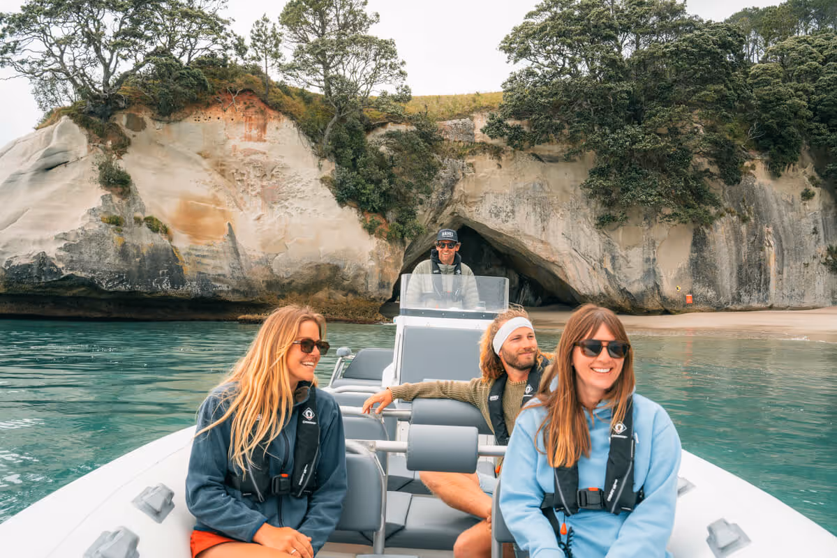 Four smiling people in casual outdoor clothing on a boat near a rocky shore with a large cave and green trees.