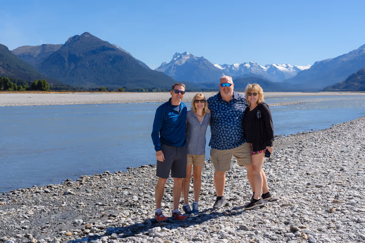 Four adults wearing sunglasses standing close together on rocky riverbank with mountains and clear blue sky in the background.