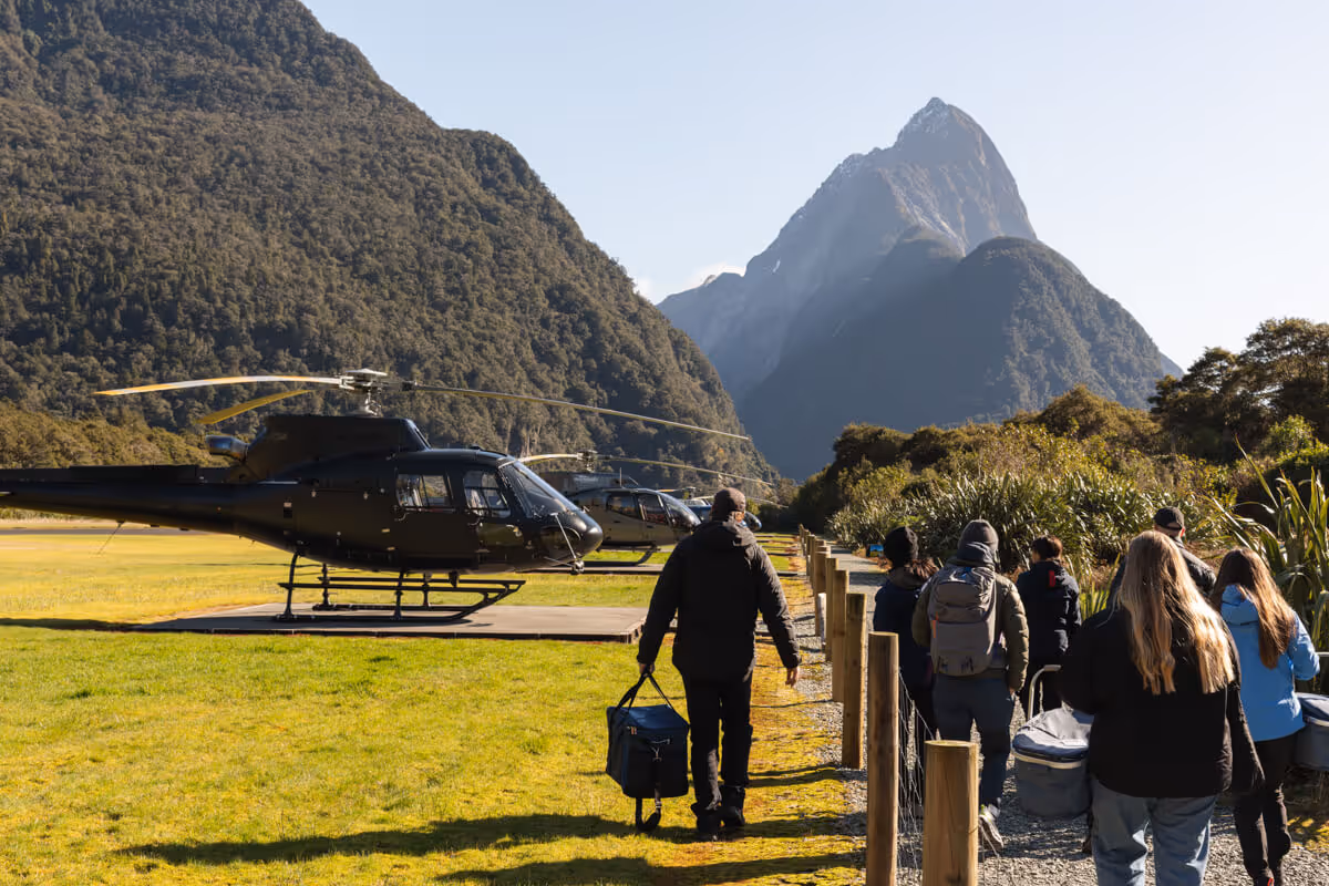 Group of tourists walking on a path near parked helicopters with forested mountains and a clear sky in the background.