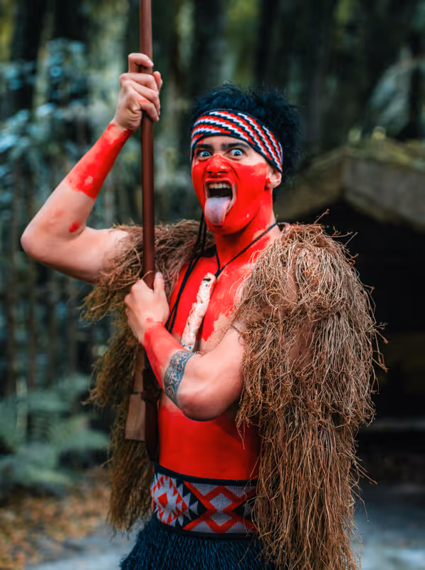 Person with red face and body paint, wearing traditional attire and a woven headband, sticking out their tongue and holding a wooden staff.