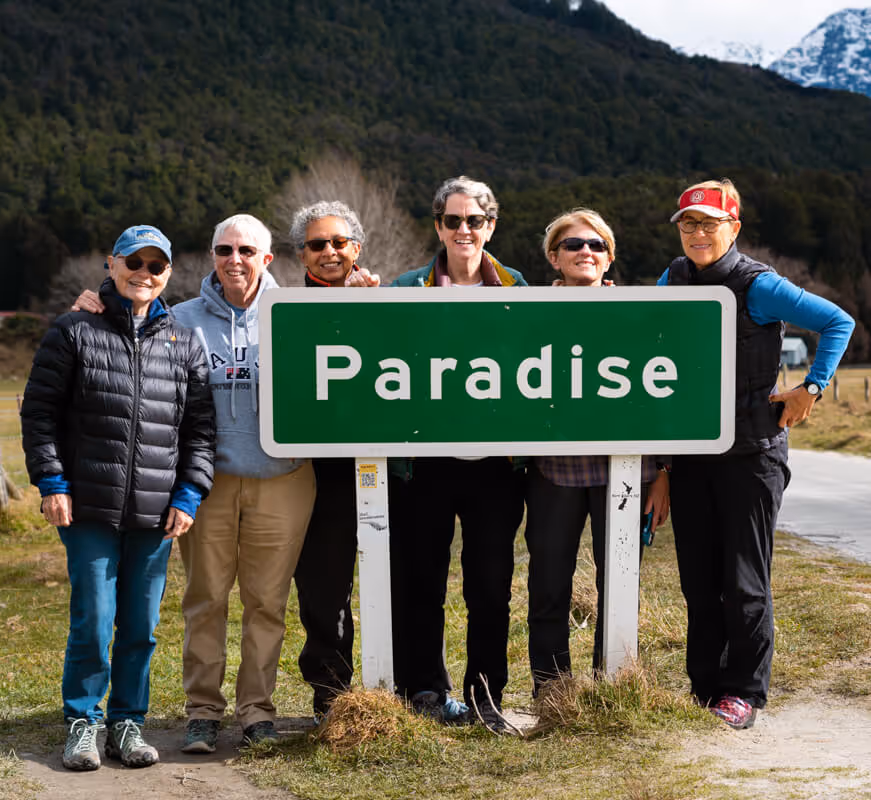 Six smiling older adults standing behind and around a green road sign that says 'Paradise' in a grassy outdoor area with mountains in the background.