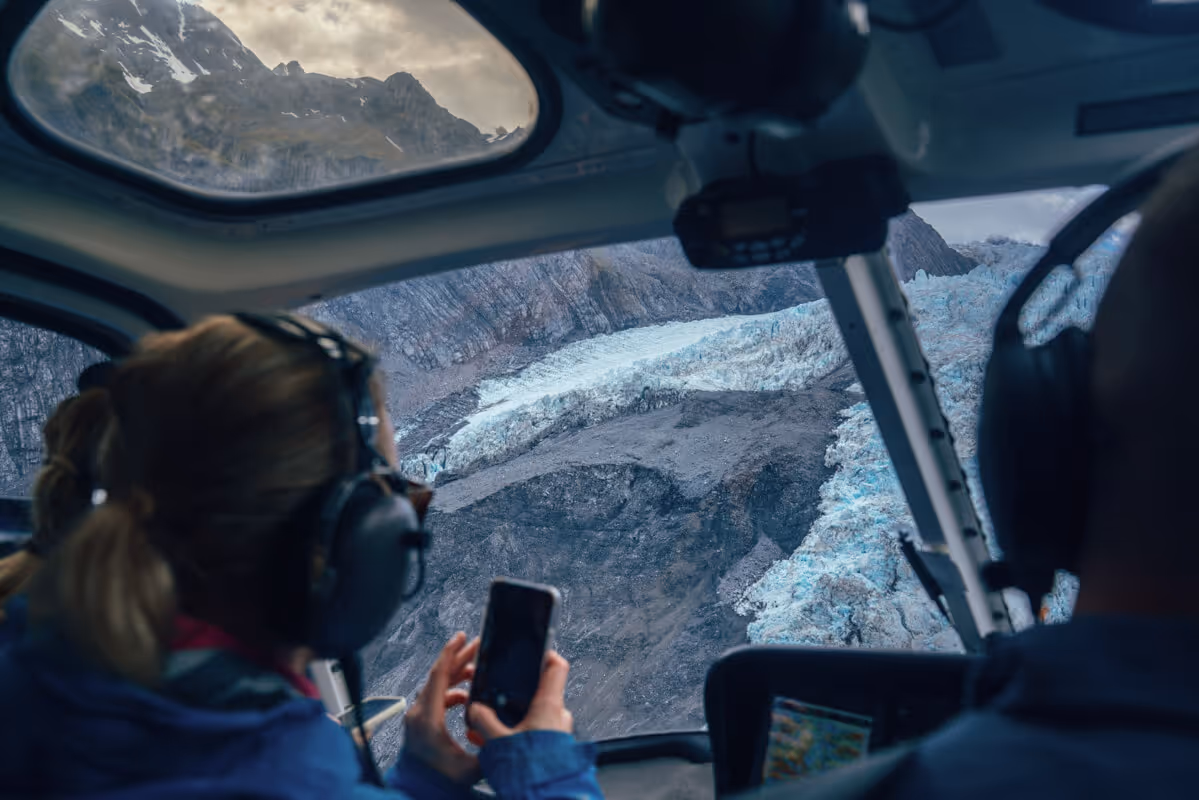 View of a glacier and rocky mountains through the cockpit window of a helicopter with two people wearing headsets inside.