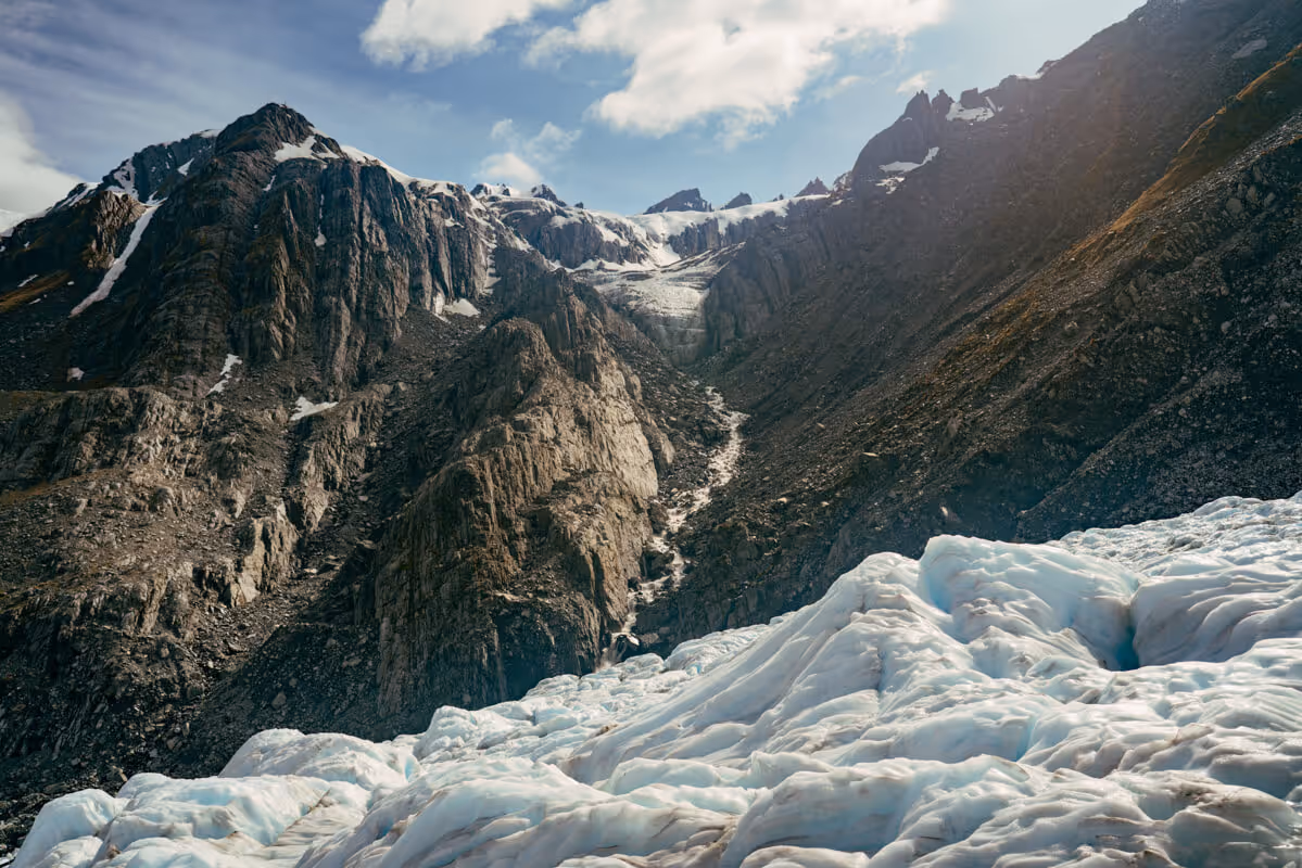 Snow-covered glacier in the foreground with rugged rocky mountains under a partly cloudy sky.