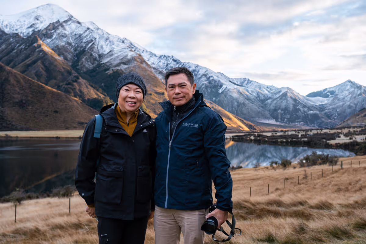 Smiling couple dressed in outdoor jackets posing in front of a lake with snow-capped mountains in the background.
