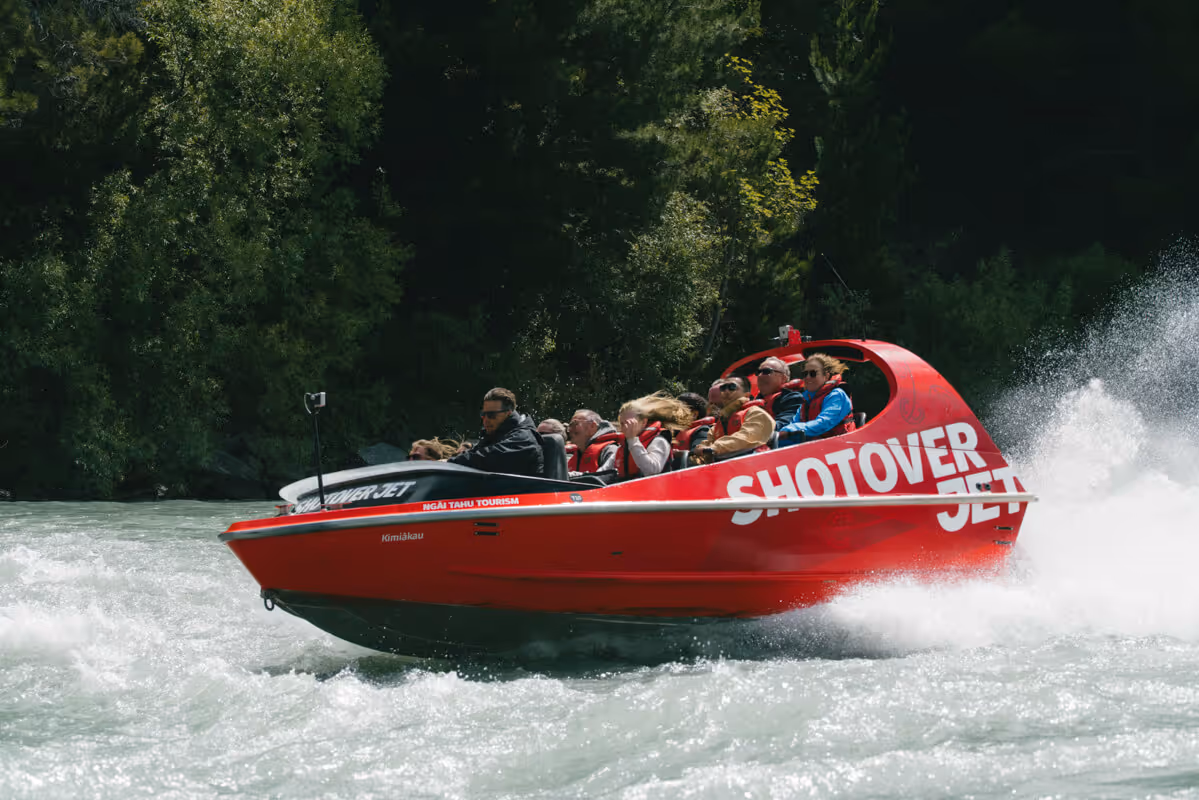 Group of people wearing life jackets riding a red Shotover Jet boat on a fast-moving river surrounded by green trees.