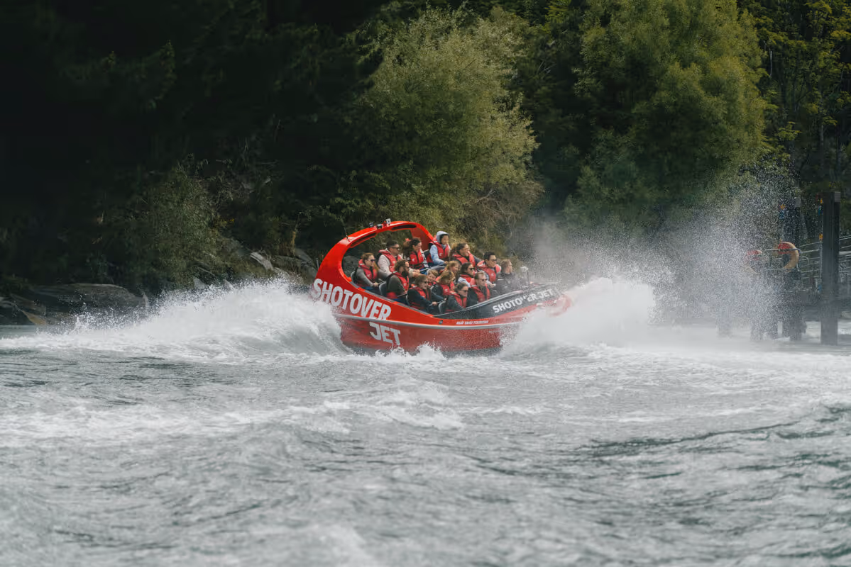 A group of people wearing life jackets on a red Shotover Jet boat speeding and splashing water on a river surrounded by dense green trees.