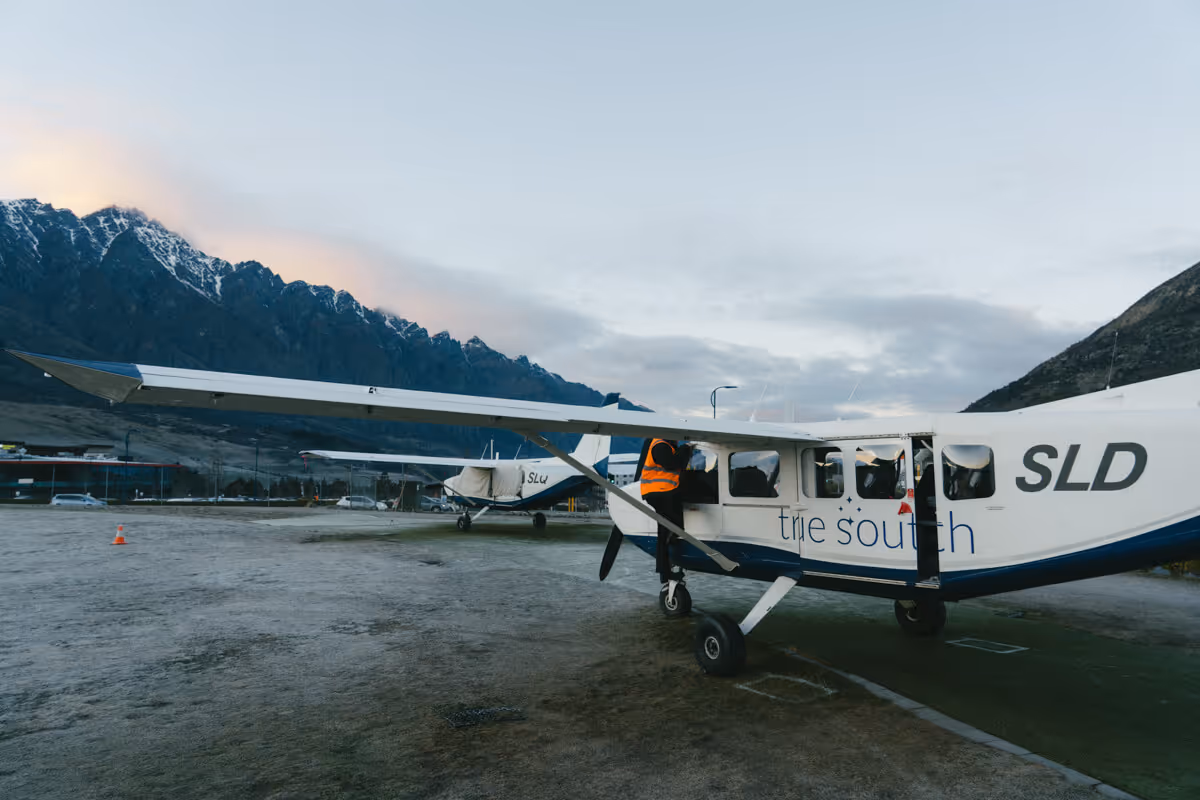 A small white and blue plane parked on a frosty airfield with mountains in the background, and a person in an orange vest near the door.