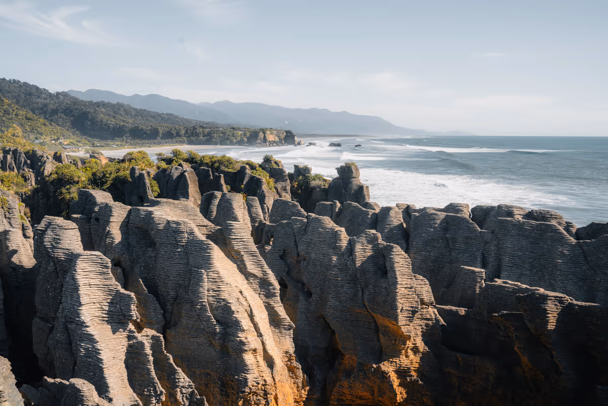 Pancake-shaped layered rock formations along a coastline with ocean waves and forested hills in the background.
