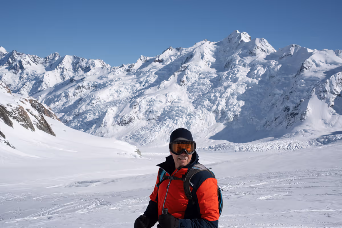 Man in red and black winter jacket with ski goggles standing on snowy mountain slope with snow-covered peaks in the background.