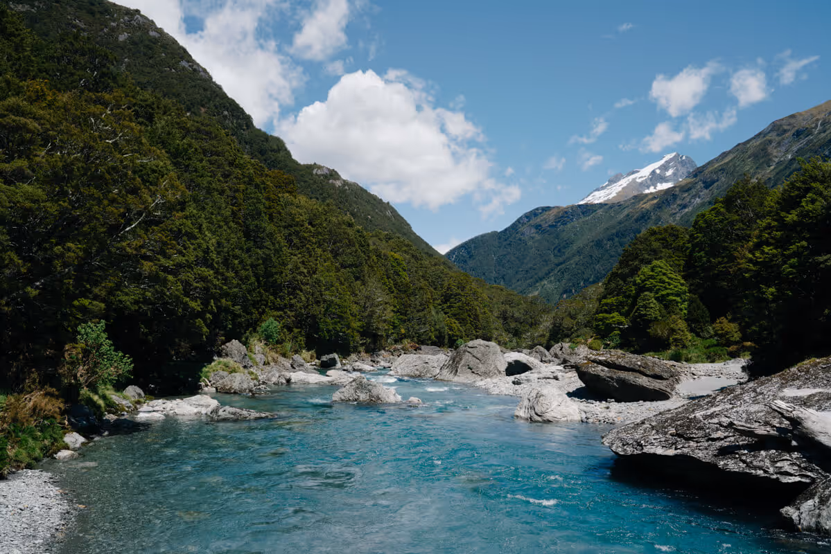 Clear blue river flowing through a rocky valley surrounded by dense green forest and snow-capped mountains under a partly cloudy sky.
