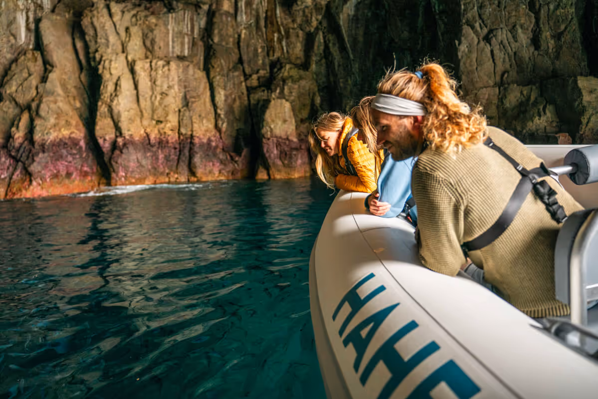 Three people lean over the edge of a small boat looking into calm water near a rocky cave wall.
