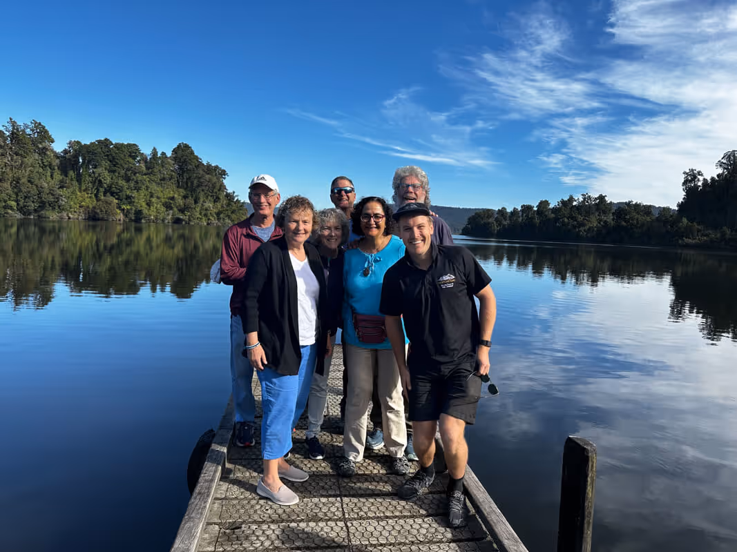 Group of seven people smiling and standing on a wooden dock by a calm lake surrounded by trees under a partly cloudy blue sky.