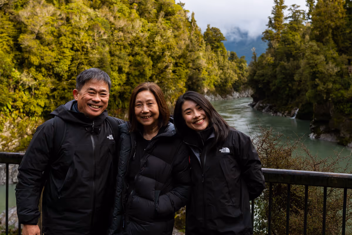 Smiling family of three wearing black jackets posing by a metal railing with a green river and lush forest in the background.