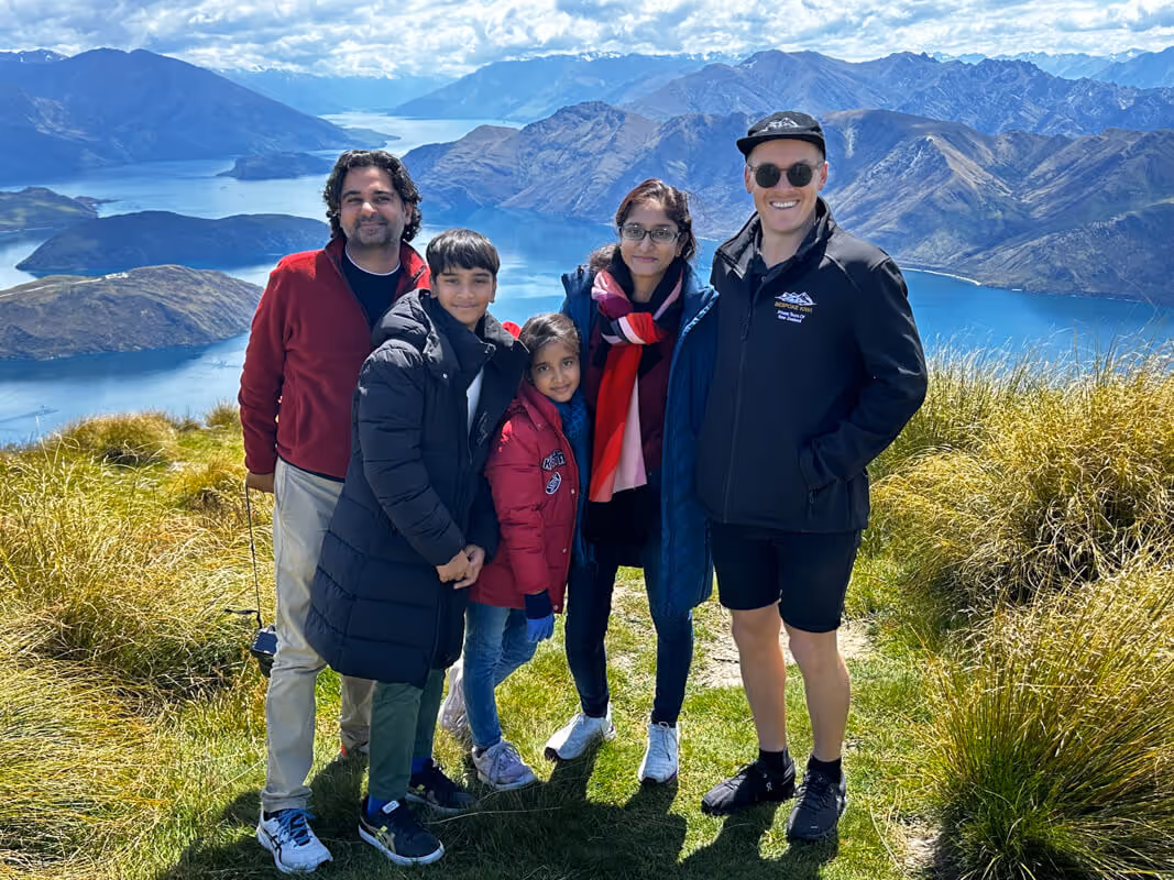 Group of five people standing on a grassy hill with a scenic lake and mountain range in the background on a sunny day.