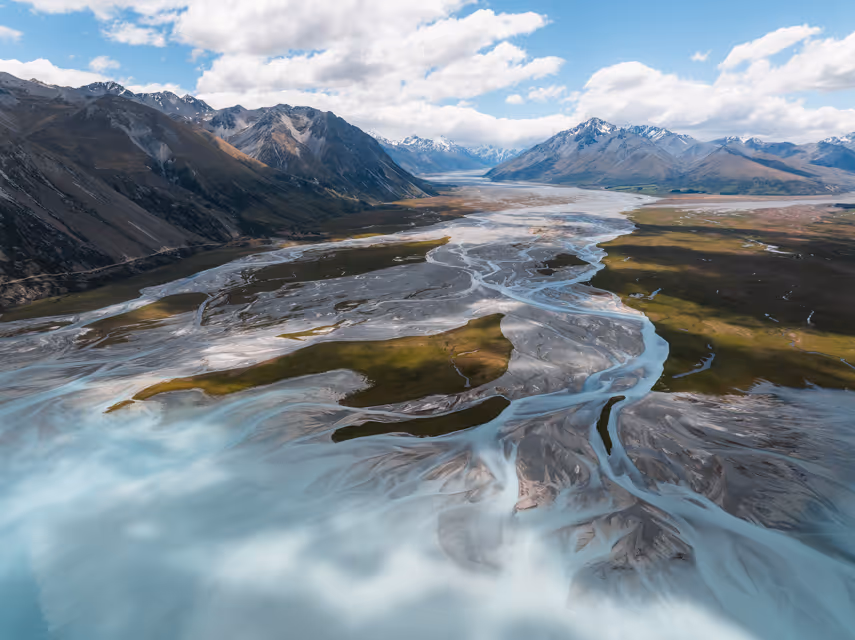 Aerial view of a braided river flowing through a valley with grassy patches and surrounded by snow-capped mountains under a cloudy sky.