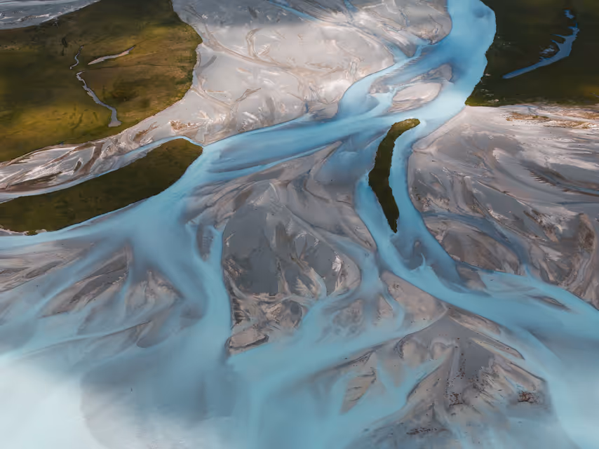 Aerial view of light blue braided river channels flowing through sandy and grassy land.