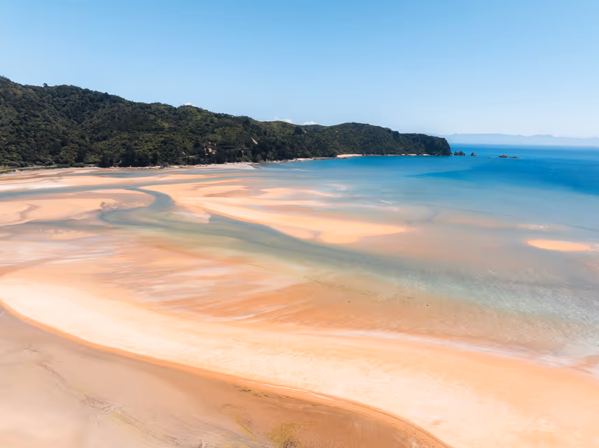 Aerial view of a sandy beach with shallow turquoise waters flowing into a bay bordered by forested hills under a clear blue sky.