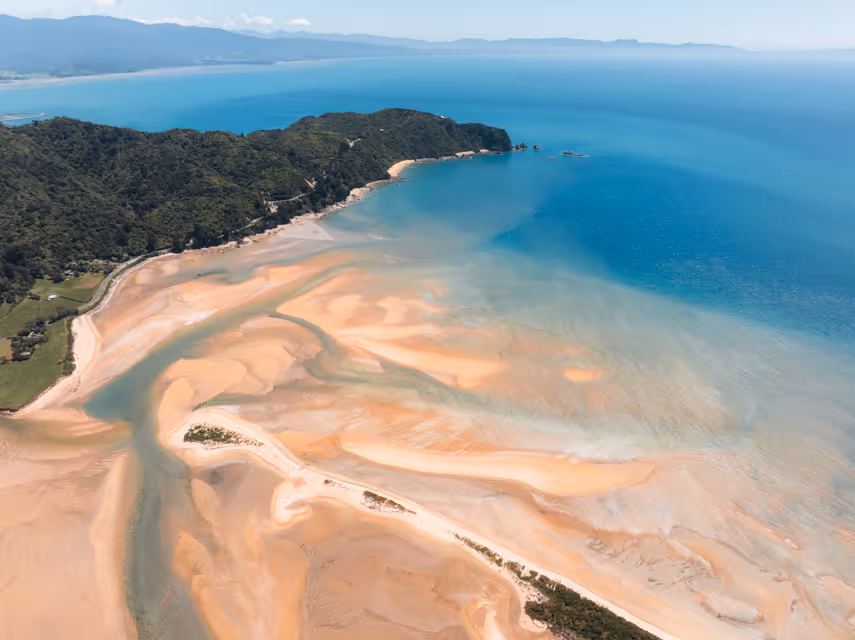 Aerial view of a sandy coastal bay with turquoise water, sandbanks, and lush green forested hills.