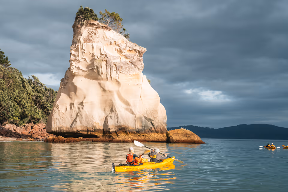 Two people kayaking near a large sunlit rock formation with trees on top, under a cloudy sky.