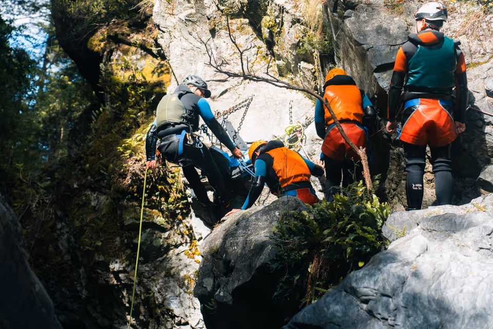 Group of people in helmets and wetsuits rappelling down a rock face in a forested canyon.