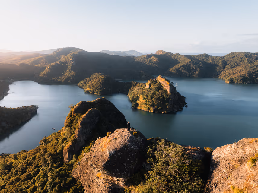 Aerial view of a person standing on a rocky cliff overlooking a lake surrounded by forested hills and small islands.