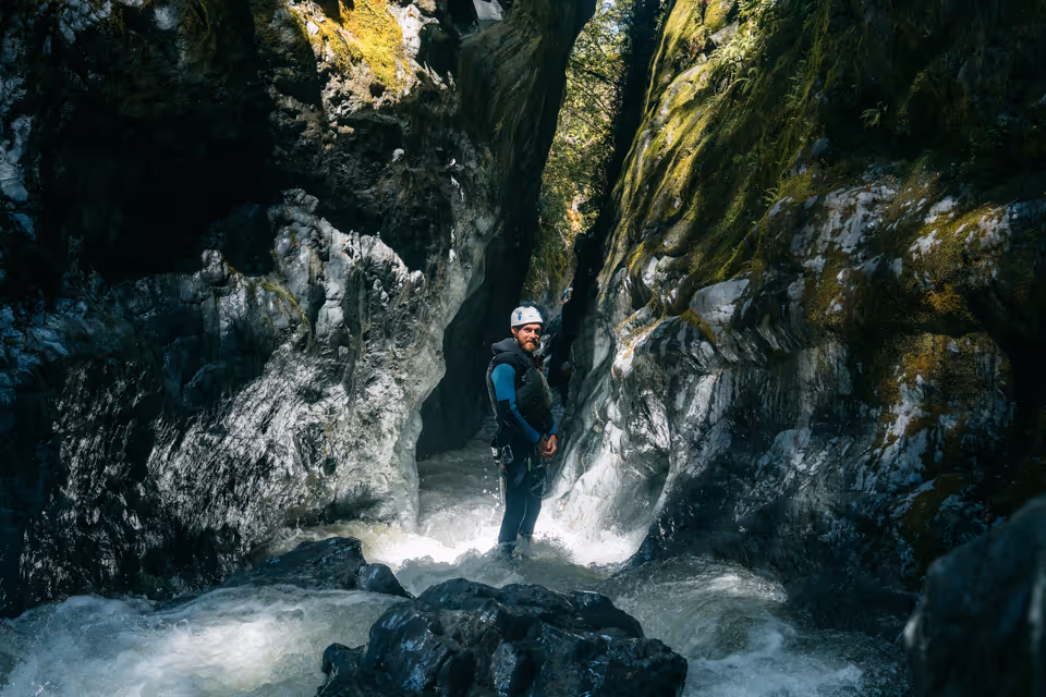 Man wearing a helmet and wetsuit standing in a narrow rocky canyon with flowing water.