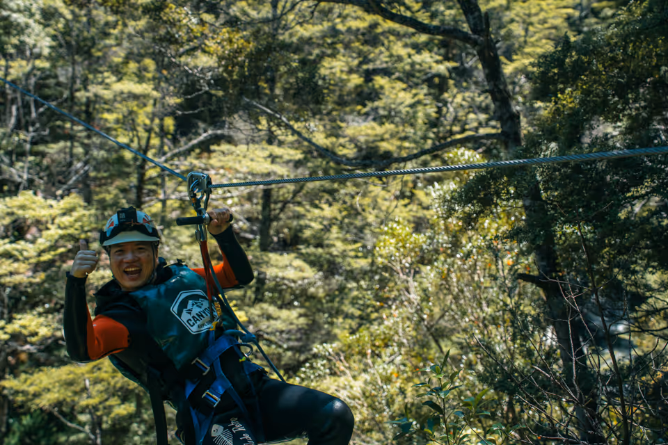 Person wearing a helmet and harness smiling and giving a thumbs-up while ziplining through a forest.