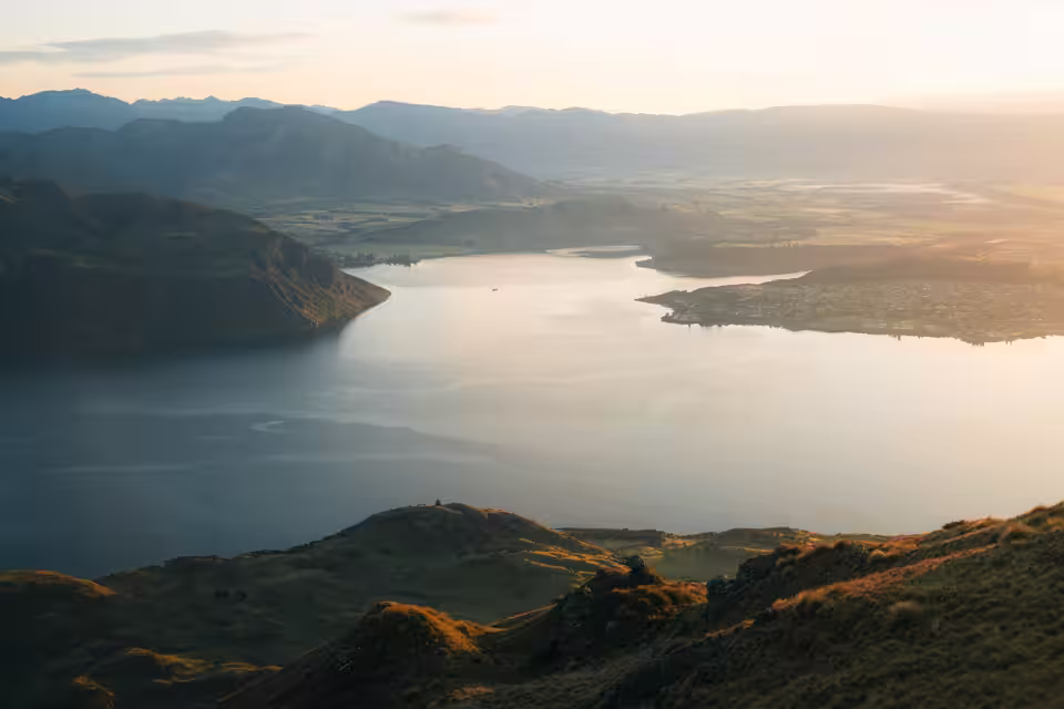 Aerial view of a large lake surrounded by green hills and mountains during sunset with soft light illuminating the landscape.