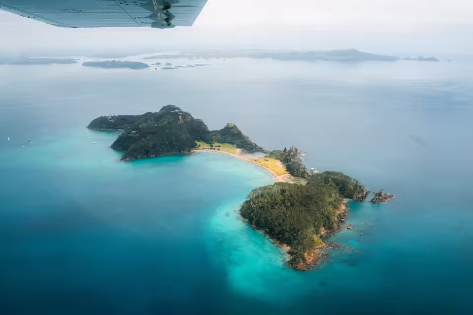 Aerial view of a small forested island surrounded by turquoise and deep blue ocean waters with a sandy beach, seen from beneath a plane wing.