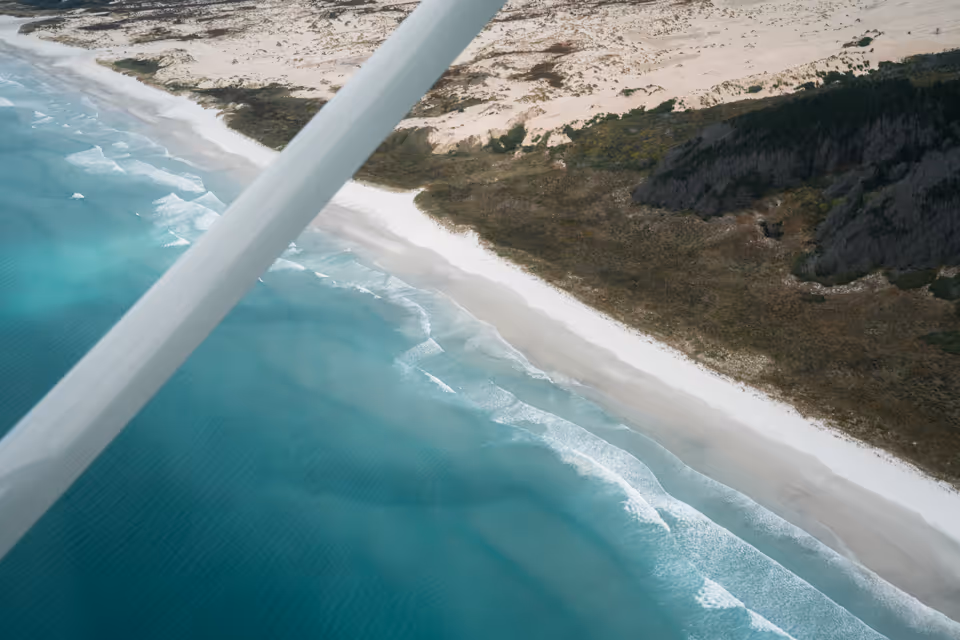 Aerial view of a turquoise ocean meeting a white sandy beach with a forested area behind it, partially obscured by a plane wing.