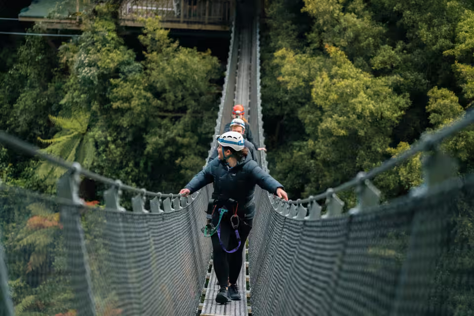 Three people wearing helmets and harnesses walking across a narrow suspension bridge surrounded by dense green forest.