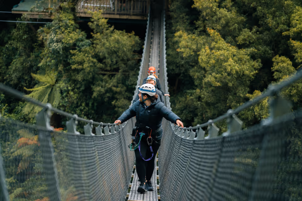 Three people wearing helmets and harnesses walking across a narrow suspension bridge surrounded by dense green forest.