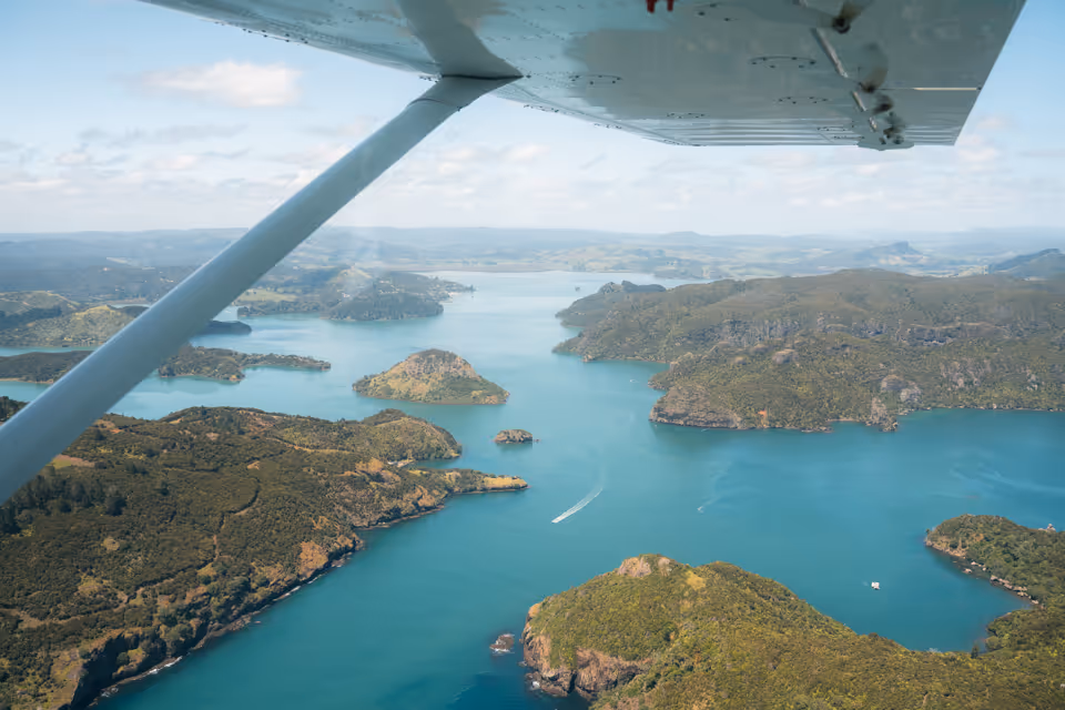 Aerial view of a large blue inlet with forested islands and a boat leaving a white wake, seen under the wing of a small plane on a partly cloudy day.
