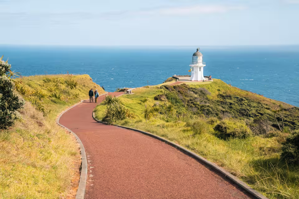 Winding pathway through green hills leading to a small white lighthouse overlooking the blue ocean.