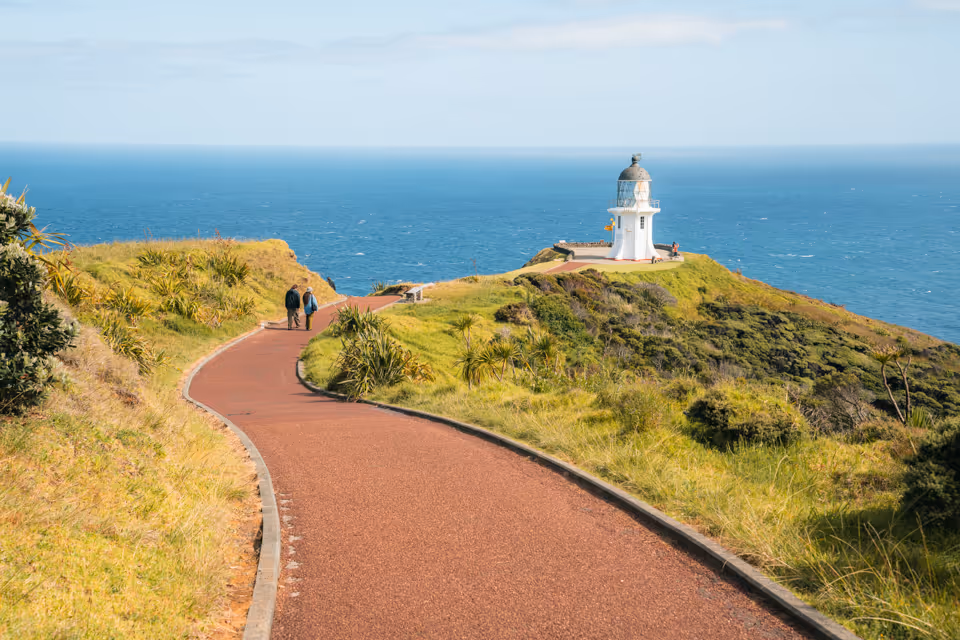 Winding pathway through green hills leading to a small white lighthouse overlooking the blue ocean.