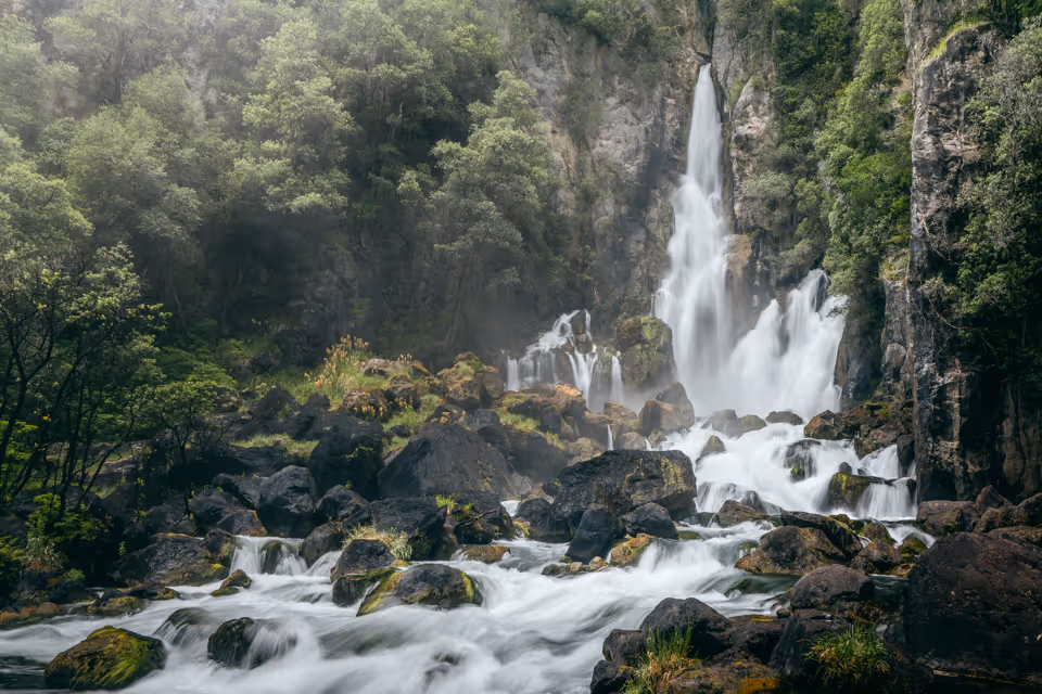 Multi-tiered waterfall cascading down rocky cliffs surrounded by dense green forest and flowing into a river with moss-covered rocks.