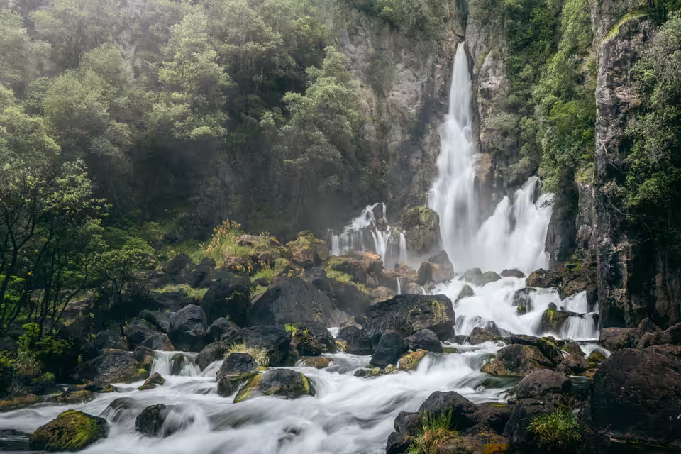 Multi-tiered waterfall cascading down rocky cliffs surrounded by dense green forest and flowing into a river with moss-covered rocks.