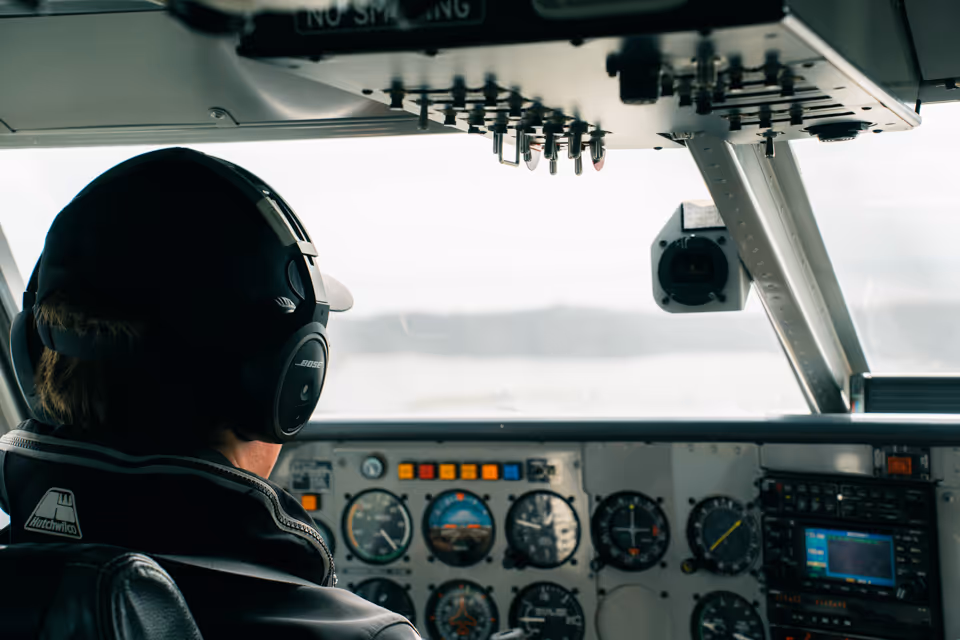Pilot wearing a hat and Bose headphones sitting in an aircraft cockpit with instrument panels and controls visible.