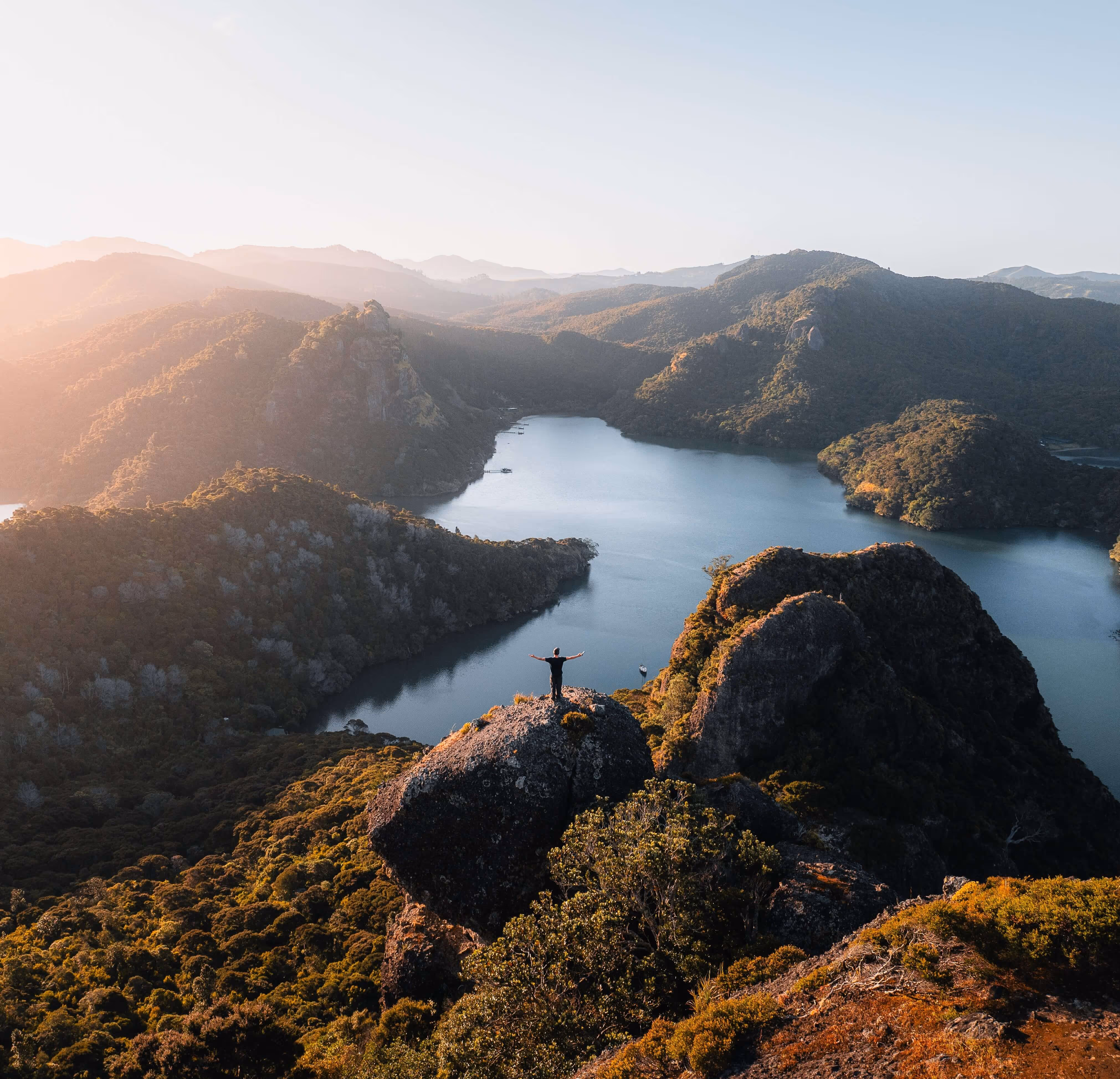 Person standing with arms outstretched on a large rock overlooking a lake surrounded by forested hills at sunset.