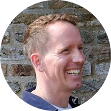 Andrew Dicker smiling with short dark hair, wearing a blue shirt in front of an exposed brick wall.