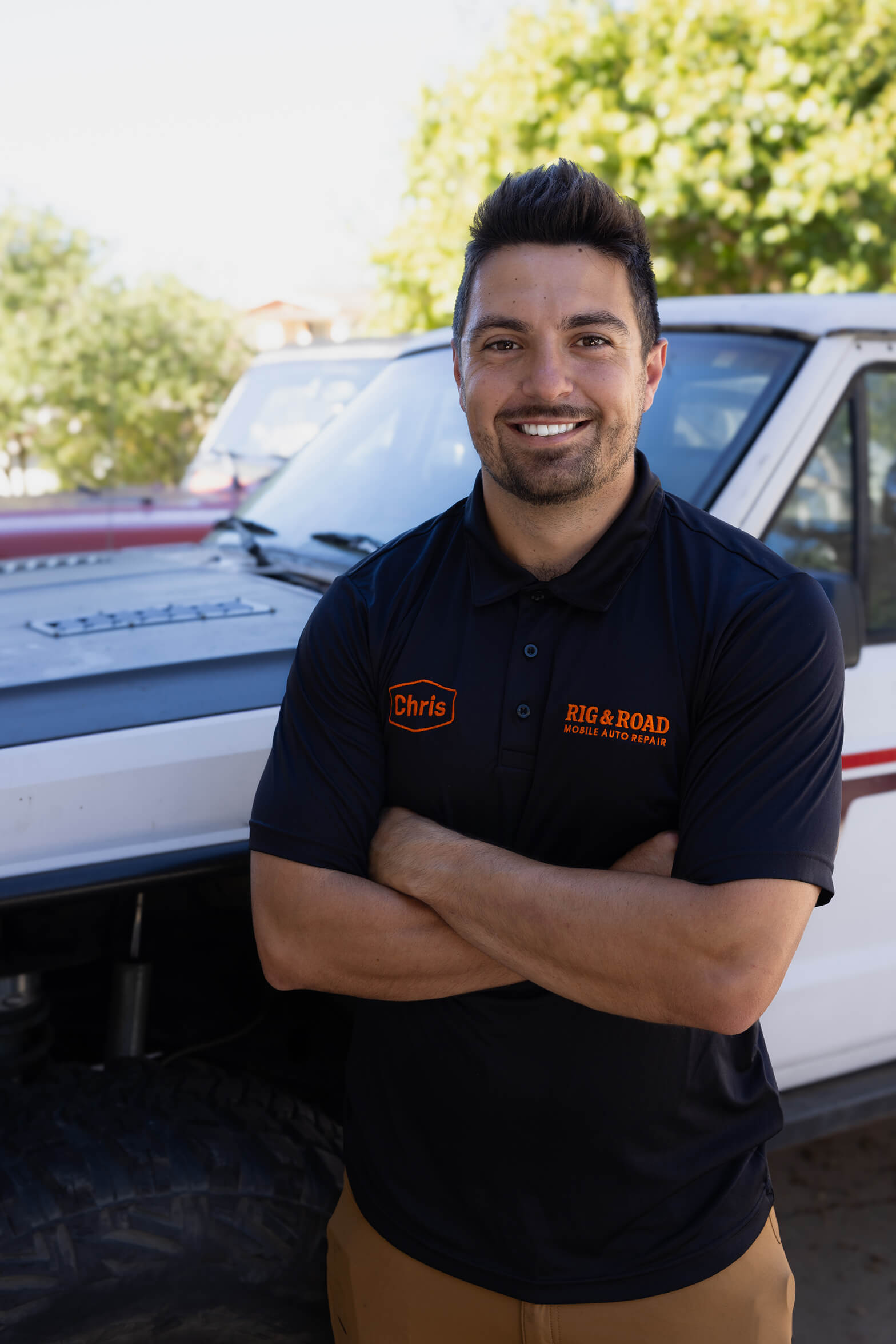 Man with crossed arms smiling in front of a white off-road vehicle, wearing a black polo shirt with 'Chris' and 'RIG & ROAD Mobile Auto Repair' logos.