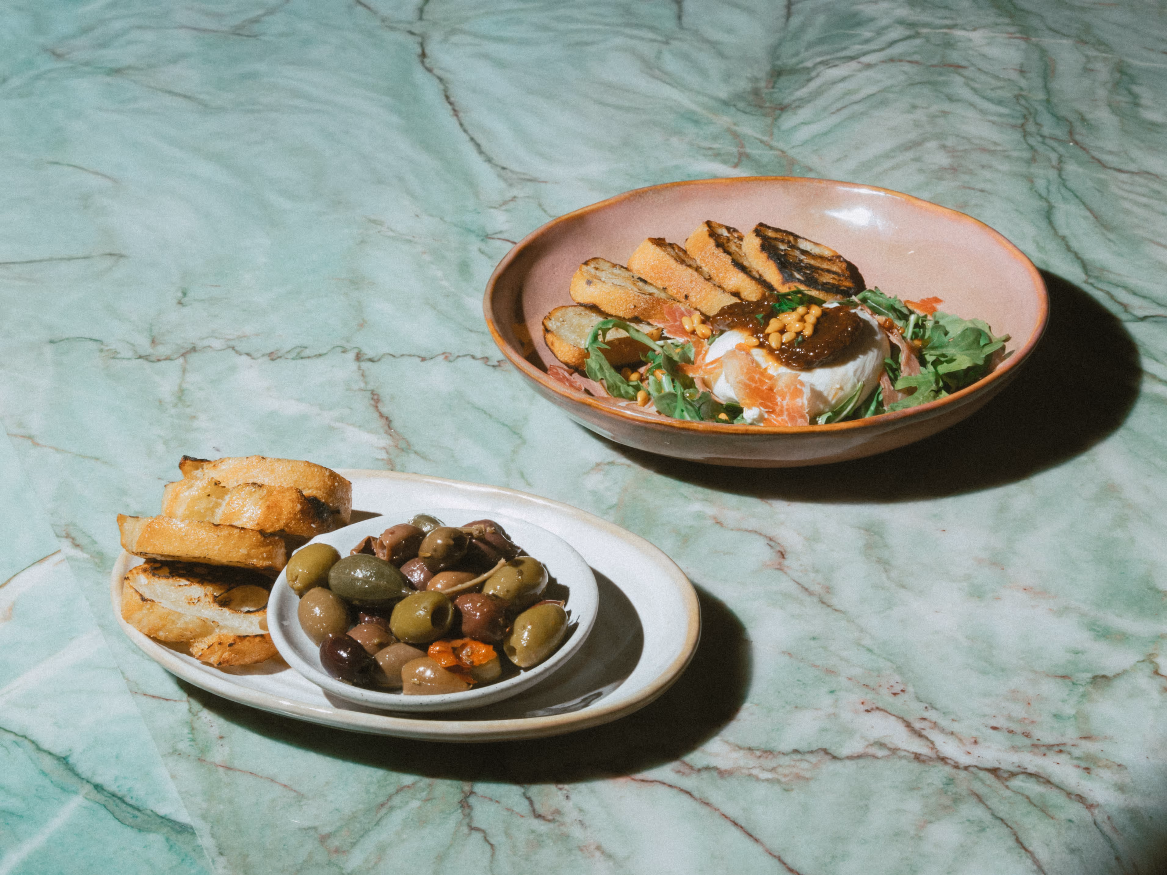 Two plates on green marble surface; one with assorted olives and grilled bread, the other with burrata cheese, arugula, pine nuts, and grilled bread slices.