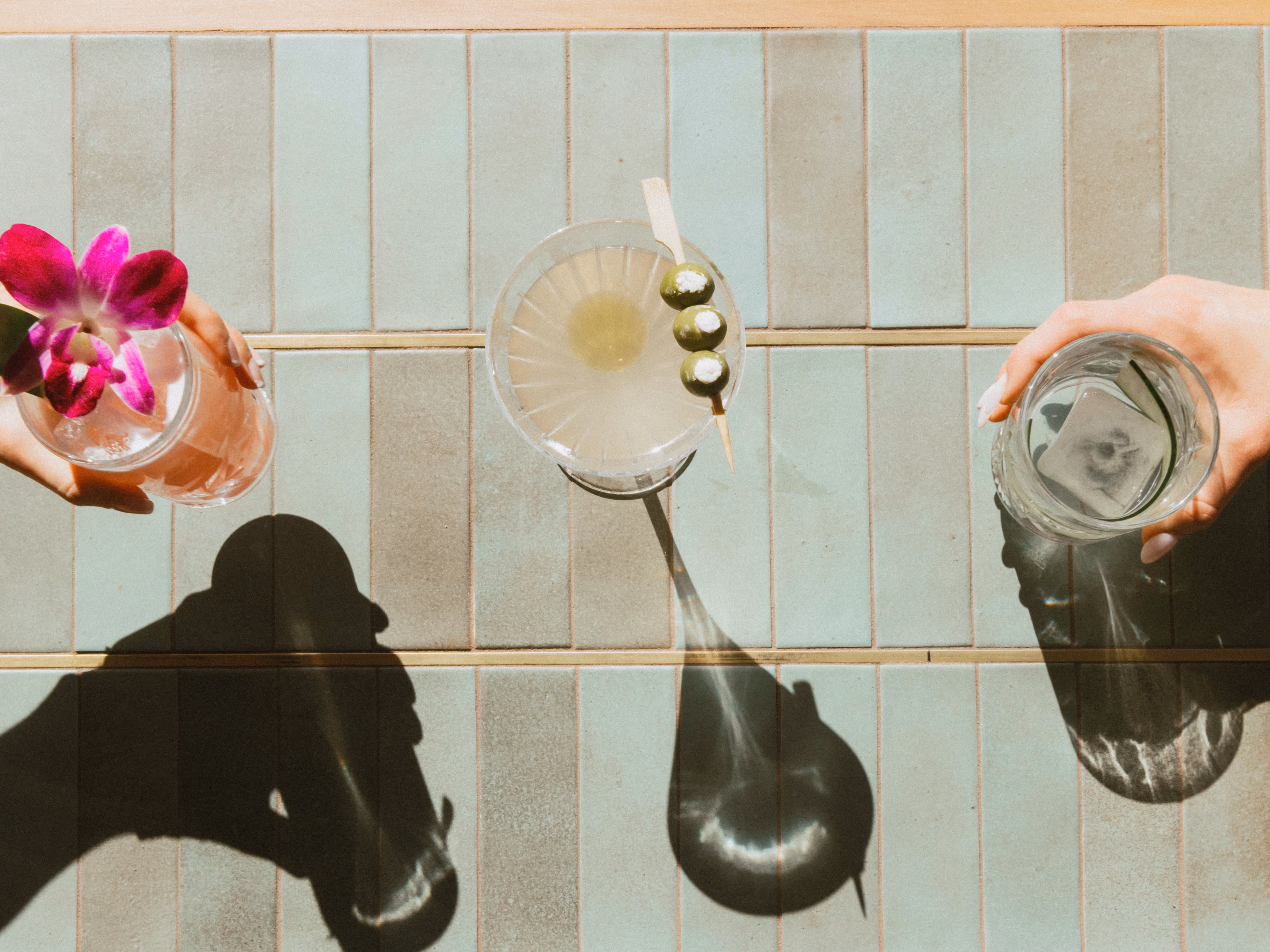 Overhead view of two hands holding cocktails on a tiled surface, one with a pink flower garnish and the other with a cucumber slice, alongside a martini glass with three olives on a skewer.