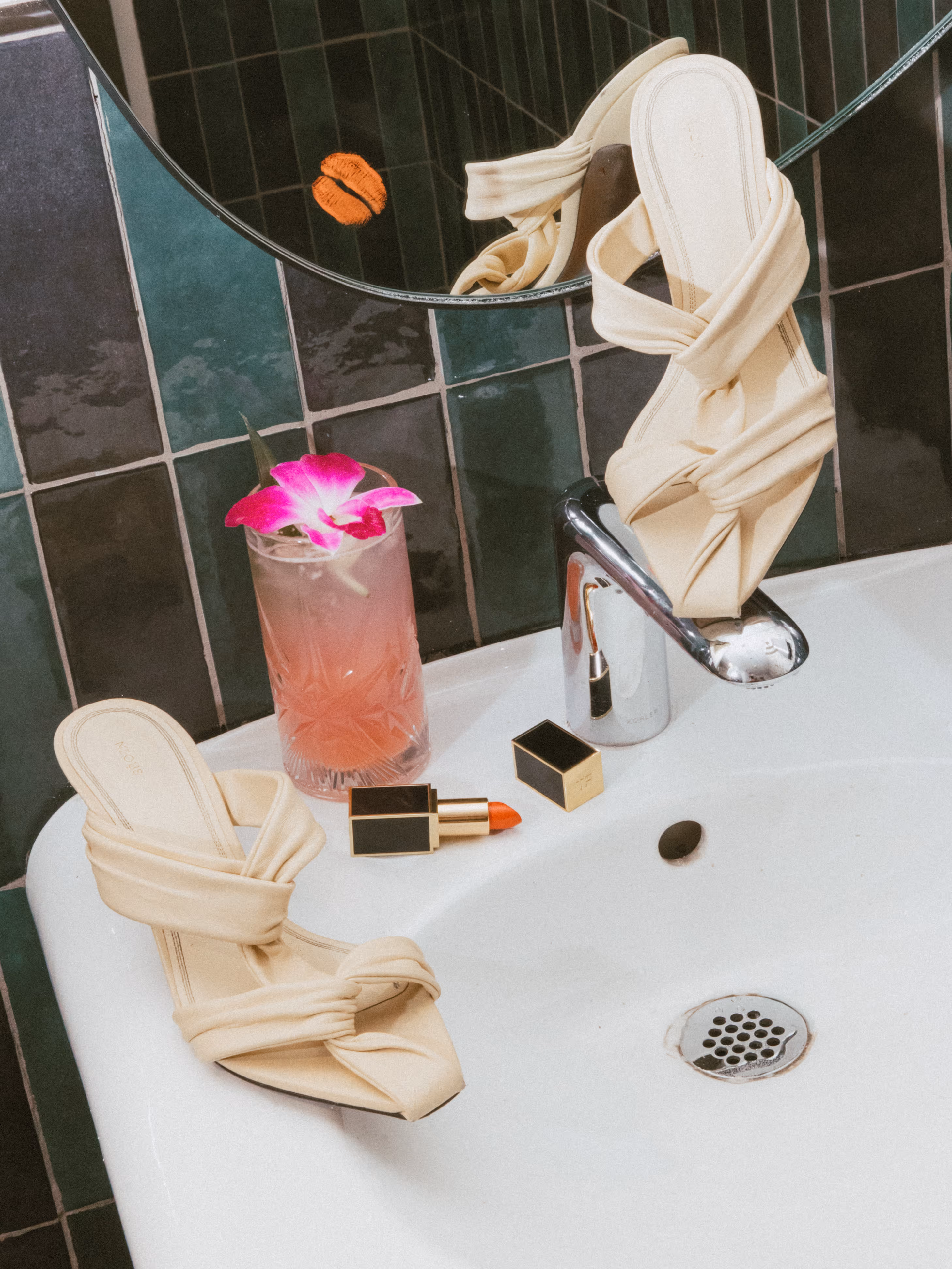 Beige strappy heeled sandals resting on a white bathroom sink with a pink cocktail garnished with a purple flower and an open tube of red lipstick nearby.