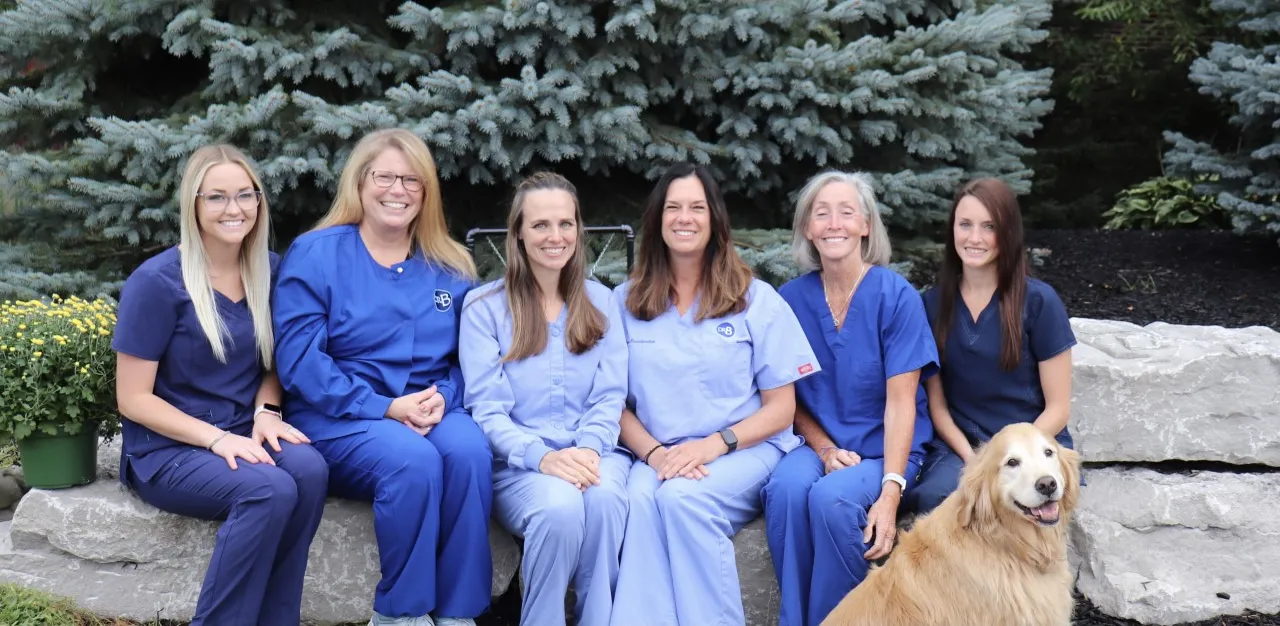 Group photo of the Boeschenstein & Middleton Family Dentistry team sitting outdoors on stone steps, smiling together in blue and navy scrubs with a golden retriever in front, surrounded by green shrubs