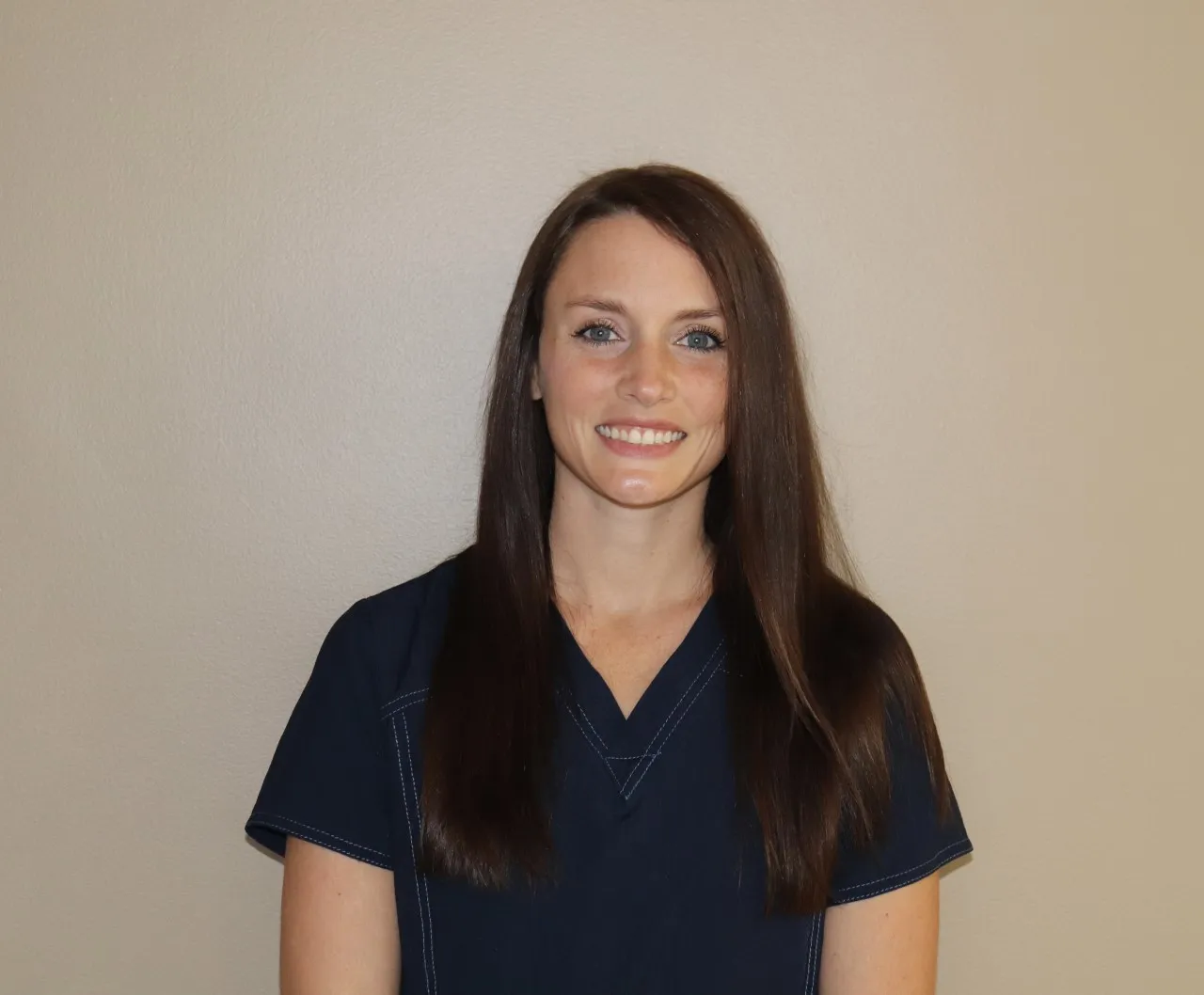Brunette dental hygienist with straight hair wearing navy scrubs, smiling confidently in front of a beige background