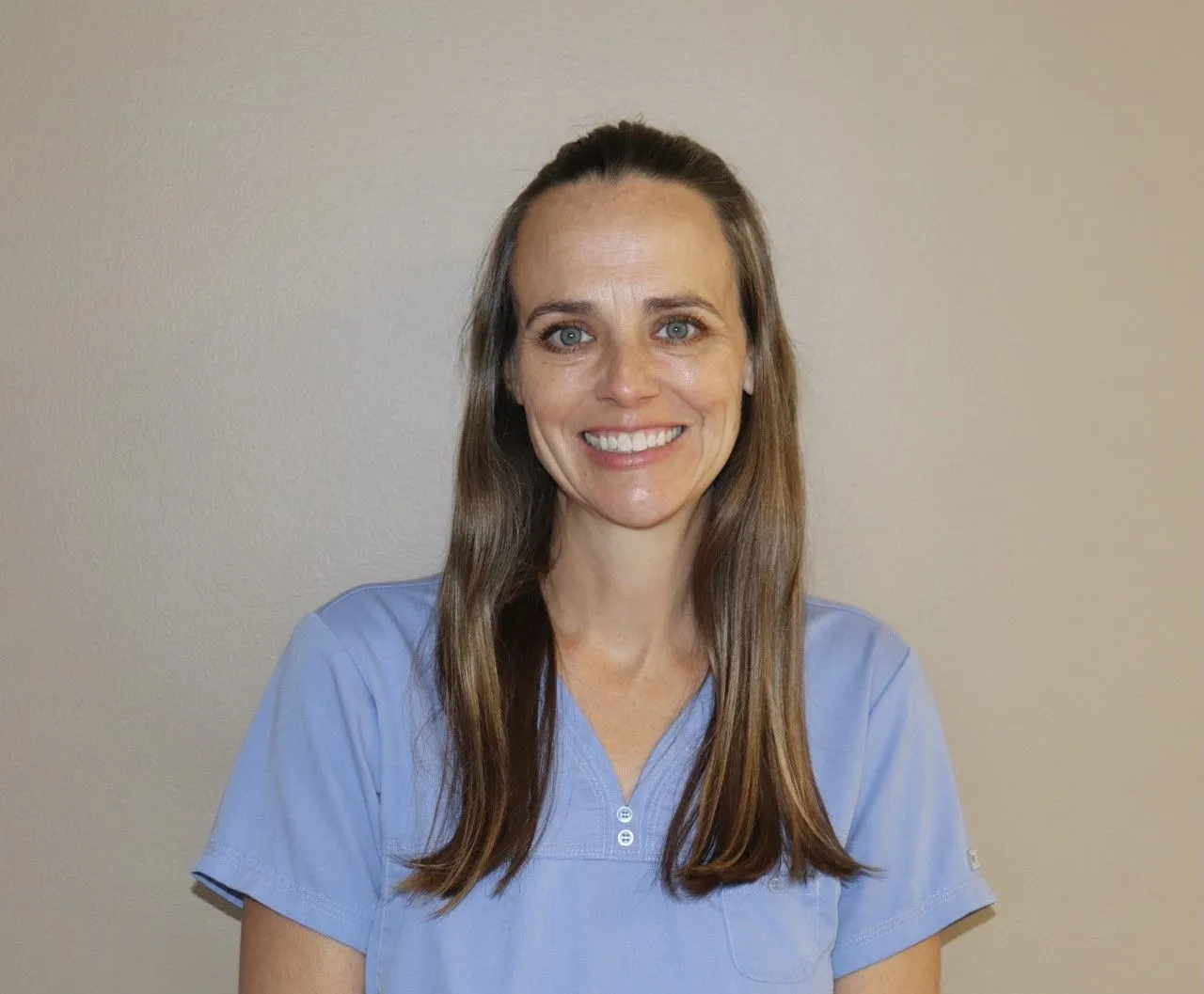 Female dental assistant with long brown hair tied back, wearing light blue scrubs, smiling brightly in front of a beige background