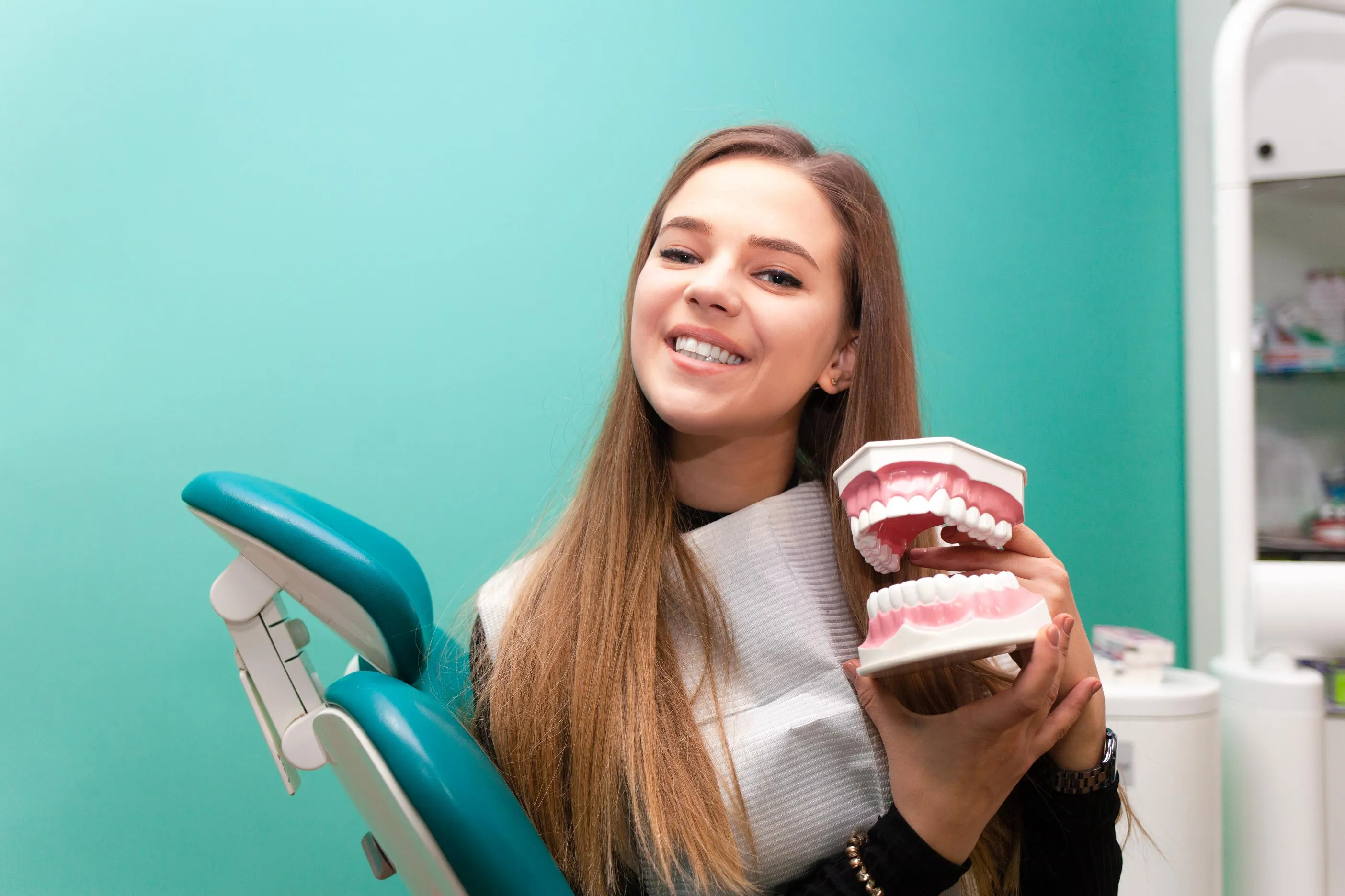 Smiling woman sitting in a dental chair holding a model of teeth while demonstrating proper oral hygiene and preventive dental care in a modern clinic